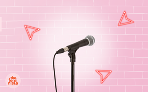 Stand-up comedy microphone in a spotlight on a pink brick background with neon arrow and The Feminista logo
