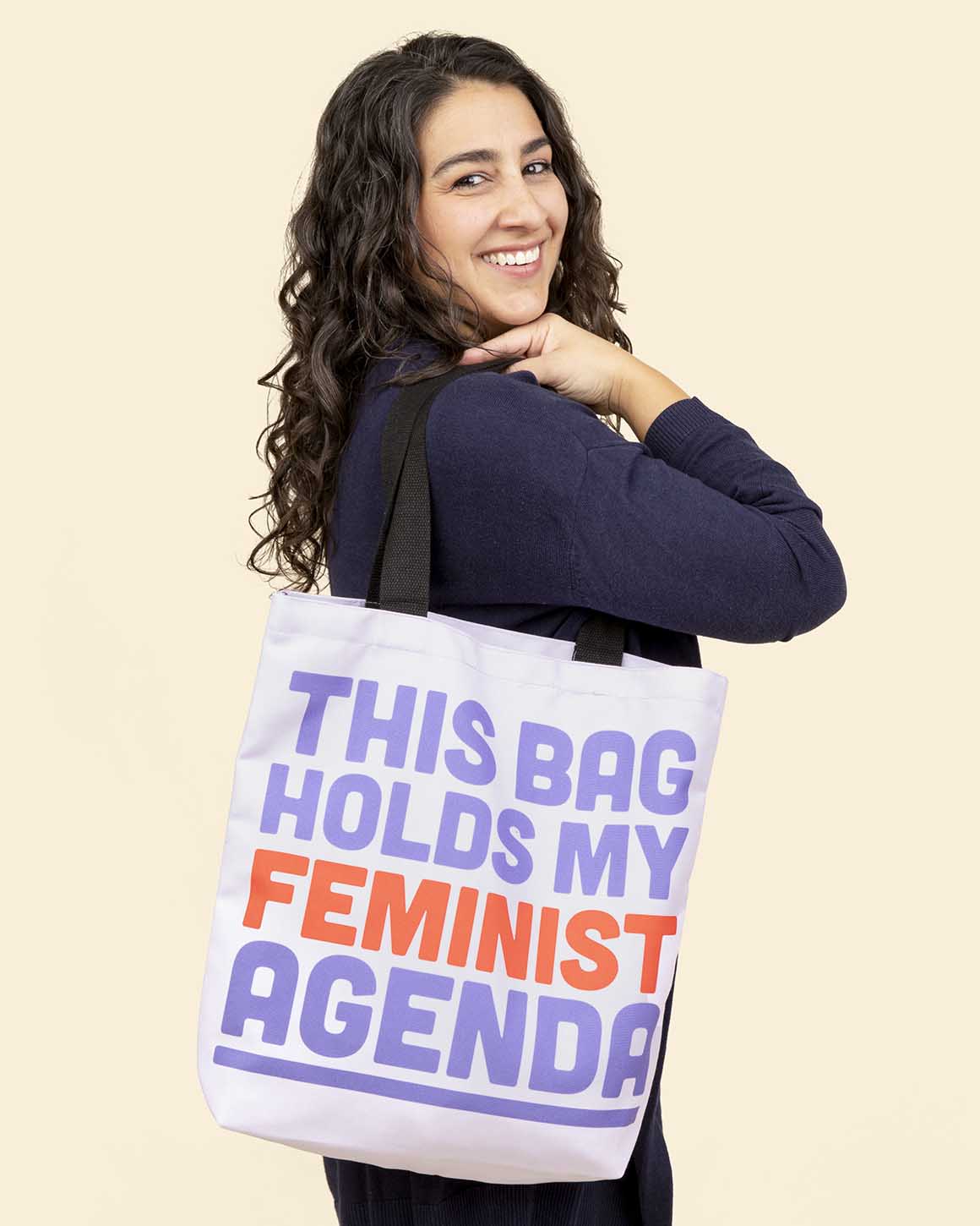 Smiling woman holding a bold purple tote bag that reads this bag holds my feminist agenda